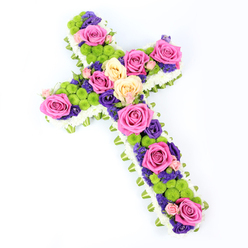 Photographed against a flawless white background, this floral cross appears as a serene, carefully shaped tribute, angled gently from the upper left to the lower right to show its dimensionality. The border of the cross is crafted from a dense ring of white spray chrysanthemums, the tiny blooms packed so closely that they form a plush, cloud-like frame. Just beneath this crisp white outline, a ribbon of pale green-either folded satin or neat foliage-traces the full shape, introducing a soft, fresh accent reminiscent of the gentle greens around Mill Hill Park. Within this delicately layered frame, the centre of the cross is richly planted with an array of blooms. Dominating the design are generous roses in hot pink and creamy pale yellow tones, their full, open heads revealing intricate swirls of velvety petals. Between them nestle clusters of lime-green button chrysanthemums, their compact, spherical forms offering contrast and a pop of modern brightness. Scattered through the spaces are small blossoms in deep purple and violet, such as statice or lisianthus, which add depth and a sense of quiet contemplation. The flowers are arranged in a way that guides the gaze slowly along the length of the cross, inviting reflection. The bright, even lighting highlights every texture-from the smooth rose petals to the slightly tufted centres of the green chrysanthemums-while the white background keeps the focus firmly on the tribute itself. This floral cross, crafted with care by experienced florists serving the Mill Hill area, would be a fitting expression of sympathy and faith for a funeral, wake, or memorial gathering, offering a gentle blend of remembrance and hope.