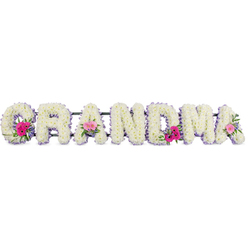 In this image, the name "GRANDMA" is beautifully rendered in flowers, each bold letter composed of tightly packed white chrysanthemums that give the tribute a full, plush appearance. The blooms are arranged in a slightly domed style, so the surface of the letters looks softly rounded rather than flat, suggesting warmth and comfort. Every letter is framed by a slender border of lilac ribbon, neatly pinned along the outer edge. The satin ribbon adds a gentle, pastel glow that traces the outline of the word and subtly softens the transition between the white flowers and the background. At three considered points in the arrangement, the florist has introduced accent posies. One sits on the sweeping curve of the "G", another on a central "A", and the third rests on the final "A" at the end of the word. These small bouquets combine lively pink gerberas, delicate pink roses, and sprigs of fresh greenery, giving the tribute touches of cheerful colour without overwhelming its calm, reflective character. The flowers look freshly conditioned, as though arranged by hand in a Mill Hill workshop near the quiet residential streets around Mill Hill Broadway, ready for delivery to a nearby church or cemetery. Soft, even lighting illuminates the composition, revealing the fine textures of each chrysanthemum petal and the smooth sheen of the ribbon trim. The overall mood is gentle and respectful, ideal for families across Mill Hill who wish to honour a grandmother with something both personal and understated, spelling out her role at the heart of the family in clear, graceful letters made of blooms.