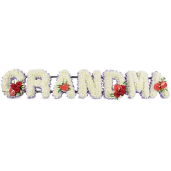 In this tender sympathy arrangement, the word GRANDMA is spelled in large, three-dimensional letters crafted entirely from fresh white blooms, evoking the quiet dignity of a funeral service in Mill Hill or a memorial gathering at a nearby church. Each capital letter is meticulously formed from densely packed white cushion chrysanthemums or similar small-headed flowers, their petals overlapping to create a smooth, plush texture that reads as soft and comforting. The outline of every letter is gently defined by a narrow border of pale purple ribbon or backing, which peeks out like a soft lilac shadow along the curves and edges and helps each character stand out clearly against the bright white background. Vivid red accents are arranged with purpose and care. On the first letter, G, a small spray of deep crimson roses is nestled neatly into the lower curve, alongside an elegant red-and-white variegated calla lily whose curved funnel shape adds a sculptural feel. The initial A is highlighted by a single, heart-shaped red anthurium at its base, its smooth, lacquered surface almost glowing. The central M is crowned with another grouping of red roses and a matching red anthurium, creating a focal point at the heart of the tribute, while the final A repeats the motif with one more solitary red anthurium. A dark green stand or frame discreetly supports the entire word, giving the impression of stability and continuity when seen from the front. The overall mood is one of reverence, warm memories, and gentle farewell, as though a family in Mill Hill has chosen a floral tribute that speaks their love for a much-adored grandmother without needing any words spoken aloud.