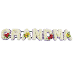 A beautifully structured GRANDMA floral tribute fills the frame, crafted with the care and attention you would expect from an experienced Mill Hill florist preparing flowers for a family farewell. The seven letters stand in a clean horizontal line against a bright, uncluttered white backdrop, each one clearly defined and easy to read. Every letter is packed tightly with small white chrysanthemums, or similar button-shaped blooms, so that their petals overlap and create a smooth, padded surface with no visible gaps. The flowers appear fresh and crisp, their whiteness suggesting purity, peace, and gentle remembrance. Around the base of each letter runs a thin edging of pale lilac or soft purple flowers, forming a delicate underline that separates the letters from the background and introduces a soothing hint of colour, much like distant heather on the outskirts of Mill Hill. The design is lifted by carefully placed accent blooms. On the lower curve of the opening G, three deep red roses sit together in a compact cluster, surrounded by shiny dark green leaves, creating a rich contrast that draws the eye to the start of the tribute. The first A carries two yellow-green cymbidium orchids along its crossbar, their unusual chartreuse petals and patterned centres adding sophistication and a slightly exotic feel, perhaps reflecting the grandmother's distinctive personality. The central R, N, and D remain simple and unembellished, made only from the white flowers with the lilac trim, providing restful space in the composition. On the M, another set of three crimson red roses with verdant foliage nestles into the lower right, echoing the roses on the G and bringing visual balance. The final A repeats the pair of yellow-green orchids, gently tying the design together. All of the letters are supported by a discrete dark metal frame, almost invisible from the front, ready to be displayed at a chapel of rest, crematorium, or church service around Mill Hill. The overall impression is one of tenderness and respect, a floral tribute that expresses love and gratitude for a beloved grandmother's life.