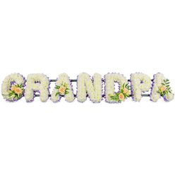 The image shows an elegant GRANDPA funeral tribute, arranged as a long row of freestanding floral letters that seem ready to be placed beside a coffin or at the front of a service in Mill Hill. Each letter-G, R, A, N, D, P, and the final A-is completely filled with tightly packed white chrysanthemums or similar small, daisy-like blooms. The flowers sit closely together, forming a plush, cloud-soft surface where their tiny yellow-green centres are barely visible, lending the piece a calm, pure look. Around the perimeter of every letter, a narrow ring of soft lavender or lilac flowers traces the outline, adding a gentle wash of colour and sharpening the silhouettes without overpowering the serene white. Thoughtful accent sprays add warmth and individuality: on the G and D, clusters of peach-toned roses, some fully open and others just starting to unfold, sit among slim white lily buds and rich green foliage, bringing a natural, garden-like touch to the tribute. Single peach roses with greenery decorate the first and last A, echoing these larger clusters and guiding the eye along the word. All the letters rest on a slim, dark support bar that runs beneath them, offering structure and stability while remaining largely hidden. Photographed against a clean white background, the textures and colours appear crisp and defined, reminiscent of walking into a quiet chapel off Mill Hill Broadway where a carefully prepared arrangement waits in stillness. The tribute gently expresses love, gratitude, and remembrance for a grandfather who meant the world to his family.