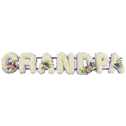 Against a clean white backdrop, the word GRANDPA is written entirely in flowers, forming a touching, three-dimensional tribute that feels deeply personal, the kind you might see at a quiet funeral service in Mill Hill. Seven capital letters stand shoulder to shoulder in a horizontal line, each one packed with a dense covering of small, ruffled white chrysanthemums or similar blooms, pressed so close that the texture appears thick and plush, almost like snow that has just settled. Encircling the edge of every letter is a delicate trim of small lilac and soft purple flowers, creating a refined outline that gently defines the shapes without overpowering the calm simplicity of the white. Inside several letters, subtle floral groupings add colour and character: the G and first A contain clusters of lily-like white flowers intertwined with bright pink and fuchsia blossoms and deeper purple notes, nestled among glossy dark green leaves. The D and one of the central As feature similar sprays, sometimes lifted further by tiny lime-green button chrysanthemums that hint at the fresh greenery of nearby Mill Hill Park. A narrow, dark green bar runs discreetly behind the letters, acting as a hidden backbone that keeps the tribute straight and strong, ready to be displayed next to a casket or memorial plaque. The lighting is soft and even, revealing every petal and curve without casting distracting shadows. Emotionally, the piece conveys a quiet mixture of sorrow and gratitude, like a final embrace for a beloved grandfather who has been at the heart of family life around Mill Hill for many years.