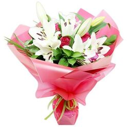 An artfully arranged hand-tied bouquet of pristine white lilies and deep red roses stands out against a clean white background, highlighting the contrast between soft petals and vibrant wrapping. At the centre, several Oriental lilies are in full bloom, their wide, trumpet-shaped flowers spreading open to display creamy white petals with gentle curves and a subtle sheen, drawing the eye towards the dark brown anthers clustered at their heart. A few lily buds, still firm and elongated in shades of green and pale cream, are interspersed among the open blooms, giving the arrangement a sense of life and unfolding beauty that will continue once it reaches a doorstep in Mill Hill. Nestled between the lilies are red roses of a rich, velvety hue, from tightly closed buds to slightly open blooms, their layered petals adding texture and a romantic depth of colour. Surrounding the central flowers is an abundant bed of foliage: broad, dark green leaves with a natural shine form a supportive base, while thinner, grass-like strands arch upward, adding lightness and movement, much like the varied greenery seen in local parks and gardens around Mill Hill East. The entire bouquet is bound together and wrapped in a bright pink, glossy cellophane or floral paper, gathered into soft, voluminous folds that ripple and catch reflections of light. Around the base, a slim lime green ribbon is tied in a neat bow, offering a crisp, playful contrast against the vivid pink. This composition, expertly crafted as if in a trusted local studio near Mill Hill Broadway, feels ideal for birthdays, anniversaries, or simply sending warm wishes and elegance to someone's home or office.
