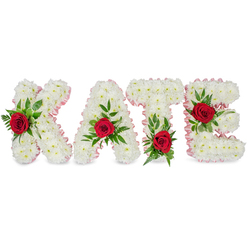 This handcrafted memorial lettering spells KATE in bold, three-dimensional block letters, each letter entirely composed of compact, pristine white chrysanthemums. The tiny petals are layered densely to create a soft, cloud-like surface that conveys purity and calm. Each letter is finished with a narrow pale pink satin ribbon folded along its edge, the subtle blush ribbon lending a refined frame to the white texture. A single, richly coloured red rose is positioned on the face of each letter as a focal accent; the roses are deep crimson and velvety, their scent faint but present in the memory of the design. Fresh, varied green foliage - broad glossy leaves paired with delicate fern fronds - cradles each rose, adding sculptural depth and a refreshing contrast of tone. Placement is deliberate: the K's rose and greenery sit on the lower section of its left vertical stroke, the A's bloom perches at the apex of its triangular peak, the T's rose settles at the base of the left part of its horizontal bar, and the E's rose is tucked into the middle horizontal arm. Presented against a pure white backdrop under soft lighting, the arrangement's textures and feminine pink edging read with clarity. Designed with the care of a Mill Hill florist, and available for thoughtful delivery across Mill Hill Village, Mill Hill Broadway and surrounding streets, this bespoke tribute is the kind of personalised funeral lettering we craft at Flowers Mill Hill for families seeking a dignified, intimate expression of remembrance.
