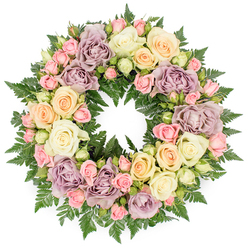 Against a clean, white backdrop, this lush circular wreath appears as a quiet, floral halo, designed as a tender farewell for a funeral or memorial in Mill Hill. The arrangement is tightly packed so that the ring is entirely covered, symbolising an unbroken bond of love and remembrance. Dominating the design are generous roses in creamy white and soft ivory, their petals fully open and layered, giving the wreath a sense of openness and honesty. Interlaced with these are roses in pale peach, delicate blush pink, and soft buttery tones, adding a gentle warmth that feels like a compassionate hand on the shoulder. Sprinkled throughout are distinctive dusty lavender and muted mauve roses, whose subtle, antique-like colour lends the composition a refined, contemplative mood. Smaller spray roses and buds are nestled between the larger blossoms, introducing delicate nuances of shape and suggesting the different stages of life. The flowers are supported by a foundation of rich, dark green fern fronds, their fine, feathery texture peeking through like the foliage in the gardens off Mill Hill Broadway and around local churches. The wreath's surface is visually rich but never chaotic; each bloom has room to breathe, and the circle remains perfectly defined. Soft, even lighting reveals the creamy tones and pastel shades without glare, enhancing the soothing effect. This handcrafted sympathy wreath, reflective of the care and detail valued by families in Mill Hill, provides a dignified, comforting tribute to honour a loved one with grace and understated beauty.