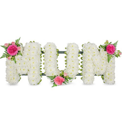 This image presents a tender memorial arrangement spelling out MUM in large, confident block letters, photographed straight on so the full shape and texture of the tribute are clearly visible. The letters rest on a dark green framework that subtly peeks through at the base, ensuring the tribute can be displayed securely beside a coffin, on a stand, or near a photograph during services in Mill Hill and neighbouring areas like Edgware or Totteridge. Every surface of the lettering is densely blanketed with small white spray chrysanthemums, their compact heads and layered petals forming a plush, almost quilted appearance. The whiteness feels calm and cleansing, with slight shadows between the blooms hinting at depth and dimension. On this pure canvas, three matching accents of vivid colour and fresh foliage bring warmth and individuality. At the upper left of the first M, a bright fuchsia garden rose unfurls, framed by softer blush-pink spray roses and partially opened buds that suggest ongoing life and memory. Fine fern fronds curve around the blooms, adding a touch of natural movement. The central U carries its own cluster at the lower middle, nestling into the letter's curve, while the upper right of the second M holds the final group of pink roses and fern. Together they form a gentle diagonal rhythm across the arrangement. The overall effect is one of quiet elegance, the kind of bespoke floral lettering often chosen by Mill Hill families to honour a mother's love at a funeral, memorial, or graveside visit.
