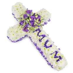 This cross-shaped sympathy tribute is tailored with intentional care, the kind of work we prepare for services across Mill Hill and surrounding Barnet streets. The main field of the cross is completely covered in compact white chrysanthemum florets, their downy texture creating a serene, cloud-like base that absorbs light softly on the white backdrop. A ruffled band of deep purple satin is woven around the perimeter, adding a dignified frame that lifts the cooler tones. The vertical arm carries the personalized message "MUM," each letter formed from smaller purple blooms - likely eustoma or spray chrysanthemums - placed densely to read clearly against the white. The centre focal spray is a luxuriant cluster of deep violet and lavender roses with pale lilac lisianthus, accented by variegated foliage whose pale edges catch light and give the bouquet depth; small green berries add a subtle, natural punctuation. The arrangement breathes a restrained elegance: the visual weight is balanced, the texture moves from pillowy white to velvety purples, and there's a soft, floral trace in the air that comforts rather than overwhelms. Prepared by Flowers Mill Hill for families in Mill Hill Village, Mill Hill Broadway and the Ridgeway, this piece is suited for wakes, graveside services, or a chapel tribute - a composed, heartfelt expression of love and remembrance.