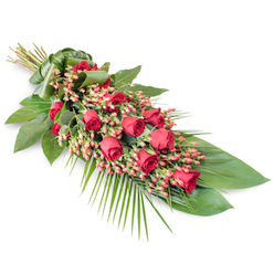On a clean, white background, this elongated sympathy spray lies at a soft diagonal, its line of flowers and foliage suggesting a serene, forward-reaching gesture. The design is anchored by a rich cluster of velvety red roses, their petals gently curving inwards and outwards in varying degrees of openness. Some roses are still in bud, symbolising lives cut short or memories still unfolding, while others are full and open, representing a life lived and cherished. Interspersed among the roses are numerous sprigs of small, round Hypericum berries in a pinkish-red hue, providing a lighter, more playful tone within the same colour family and adding a fine, bead-like texture that contrasts with the smooth rose petals. This central floral line rests upon a structured bed of deep green leaves: broad, glossy foliage gives the arrangement strength and definition, while smaller leaves and finer stems soften the edges. From the lower right edge, airy palm fronds extend in graceful arcs, creating a sense of gentle motion and elongating the form into a teardrop-like shape. At the upper left, the stems are carefully aligned and wrapped in a folded green leaf band, ensuring a tidy, understated finish that keeps the focus on the blooms rather than on visible binding. The overall lighting is soft and even, enhancing the depth of the reds and the reflective sheen of the foliage against the stark white surface. Designed with the calm, residential feel of Mill Hill in mind, near Mill Hill Park and the quiet streets off the Broadway, this tribute is ideal for funerals, cremations, or memorial services, conveying loyal love, respect, and a dignified farewell.