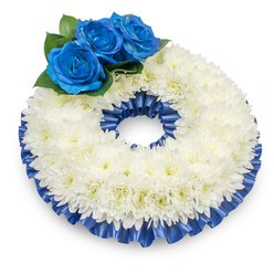 From a gentle overhead angle against a neutral white field, this circular wreath reads as a composed expression of sympathy: hundreds of compact white chrysanthemum blooms create a plush, cloud-like ring whose soft, feathery petals catch the light and form a textured, slightly domed surface. Nestled at the upper left quadrant, a trio of deep royal-blue roses rises with velvet-thick petals, each bloom carefully opened to reveal its spiralled centre; they are cushioned by glossy dark green foliage that gives depth and a living contrast to the otherwise pale ring. A finely pleated satin ribbon in matching cobalt blue trims both the inner and outer edges, the ribbon's sheen and delicate ruffling adding definition and a measured ceremonial grace. The photograph's subtle shadow beneath the wreath emphasizes the careful construction and the wreath's modest three-dimensional presence, while the overall palette-pure white and strong blue-balances tradition with personal sentiment. As an experienced local florist preparing tributes for clients across Mill Hill and Mill Hill East, I emphasise the tactile qualities: the chrysanthemums' light, papery texture against the velvety roses, the cool smoothness of the satin, and the subdued scent that suggests dignity rather than perfume. Intended for memorial services, funerals, or as a heartfelt condolence, this wreath communicates solace and respect; it would sit quietly at a service near Mill Hill Park or be delivered with care along The Broadway, offering a visual focus that honours a life with restrained beauty and thoughtful craftsmanship.