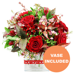 Displayed against a clean white background, this lavish red bouquet looks like it has just been arranged in a Mill Hill flower shop close to the Broadway. The eye is immediately drawn to the central cluster of luxurious red roses, their velvety petals tightly coiled in some blooms and softly unfurling in others, creating a beautiful mix of anticipation and full-blown romance. Among them, smaller deep red flowers-suggestive of alstroemeria or other fine, multi-petalled varieties-are nestled thoughtfully, adding sculptural variety and extra depth to the colour palette. Scattered delicately throughout, tiny blossoms in pale pink and white, reminiscent of waxflowers, bring a light and whimsical touch, as if tiny stars are sparkling between the larger blooms. A rich selection of green foliage frames the flowers: broad, glossy leaves provide structure and support, while finer, lighter green sprigs contribute a sense of freshness, like foliage glimpsed along Mill Hill's leafy residential streets. To deepen the mood, burgundy and reddish-brown leaves with subtle veining are artfully tucked in, echoing the rich tones of the roses and lending the arrangement a sophisticated, slightly dramatic edge. The bouquet is shaped into a rounded dome, designed so each flower has its moment yet contributes to a harmonious whole, perfect for placing at the centre of a dining table or sending as a romantic gesture. The stems stand in a modern, clear rectangular glass vase with smooth edges, where the transparent surface lets the natural lines of the stems show through. A red ribbon printed with repeating white hearts wraps around the vase, quietly reinforcing the theme of love and care. In the lower right corner, a prominent circular orange badge with bold white capital letters announces "VASE INCLUDED," indicating that this elegant, ready-to-display bouquet is ideal for Valentine's Day, anniversaries, or special celebrations throughout Mill Hill.