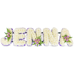This bespoke memorial arrangement reads the name JENNA in sculpted, uppercase letters formed from hundreds of creamy white chrysanthemums or similar petite blooms, each petal layered to create a pillowy, reverent texture against a plain white surface. A slender border of rich purple ribbon or tiny lavender blooms traces the inner contours of each letter, providing crisp contrast and a refined silhouette that allows the letters to sit with quiet dignity. Three small, hand-tied clusters soften the geometry: blush-pink and pale lavender spray roses, with a few creamy yellow roses and dark green fern fronds, are artfully nestled into the 'J', the middle of the first 'N', and the top right of the final 'A', introducing gentle warmth and subtle colour notes. The letters are united by slim, dark horizontal connectors that indicate a sturdy floral frame built for display at a funeral, memorial service, or celebration of life. The scent is understated-green chrysanthemum earthiness with a whisper of rose-while the texture invites a compassionate, reverent touch. Having created many such name tributes for families across Mill Hill Village and around The Ridgeway, Flowers Mill Hill understands how a personalised floral name can act as a comforting, visual tribute during grief. The arrangement is designed to be viewed from the front, where the white field and purple edging read cleanly in chapel lighting, yet it rewards a closer look with the handcrafted clusters that speak of tenderness, remembrance and enduring affection.