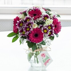 A lush, professionally arranged bouquet of fresh flowers stands centered in a clear textured glass vase, glowing against a softly diffused white background that feels like gentle daylight through a north London window. Dominating the arrangement are large gerbera daisies in deep fuchsia and magenta tones, their dark central discs creating dramatic focal points that immediately catch the eye. Around these bold faces, rounded stems of chrysanthemum-like blooms unfurl in bicolour purple and white, each flower edged in rich violet that fades towards crisp, pale tips, with sunny yellow centres that seem to echo the warmth of a late afternoon walk through Mill Hill Park. Between the stronger shades, creamy white roses in varying stages of opening - from tight buds to soft, cupped blossoms - lend a timeless elegance and suggest a gentle, powdery fragrance. Fine sprays of white baby's breath weave through the design like tiny stars, lightening the overall palette and giving the bouquet a soft, airy fullness. Deep green foliage frames and supports the flowers, adding contrast and structure while emphasising their freshness, as if they have just arrived from a local flower market near Mill Hill Broadway. The squat, jar-style vase is wrapped at the neck with a simple twine bow, adding a note of rustic charm, and a small white rectangular tag dangles from it, printed with a stylised pink rose and flowing lettering that hints at a handwritten message. The whole composition feels like a joyful, thoughtful gift - perfect for same-day delivery to a home in Mill Hill East, a welcoming reception desk, or a cosy café corner, bringing colour, celebration, and a sense of occasion into everyday life.
