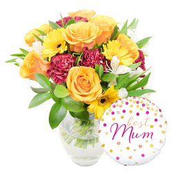 A warm, lovingly composed floral gift photographed on a pristine white background: a generous bouquet in a clear glass vase occupies the centre-left of the frame while a round, glossy white foil balloon sits to the bottom right, slightly overlapping the vase. The arrangement is anchored by full, radiant orange roses - petals unfolding in soft spirals that give off a velvety, almost peach-scented impression - paired with bright yellow gerbera daisies whose bold, flat faces and deep centres bring an immediate cheer. Rich maroon carnations, their ruffled layers dense and tactile, add a velvet depth that balances the lighter blooms, and slender white alstroemeria with faint speckling lend delicate, airy notes across the bouquet. Lush green foliage, a mix of broad glossy leaves and slender sprigs, cradles the flowers and reveals neatly cut stems through the water, a small sign of careful craftsmanship from a local Mill Hill florist. The white balloon is playfully patterned with gold and hot-pink confetti dots and tiny hearts; the centre carries the flowing hot-pink script message "best Mum," a personal, affectionate touch that makes the combination ideal for Mother's Day or a birthday tribute. Imagined on a sunny morning near Mill Hill Broadway or a quiet afternoon by the green spaces around Mill Hill Village, this gift set conveys gratitude and celebration with a fresh, lived-in charm - the kind of arrangement Flowers Mill Hill might prepare for neighbours wanting to make a beloved mum smile.