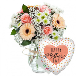 This image shows a tender, celebratory Mother's Day arrangement styled with the quiet elegance you might expect from an experienced Mill Hill florist. Against a spotless white background, a full mixed bouquet is presented in a clear glass vase, its shape simple and rounded so that the focus stays on the flowers. Around the neck of the vase, a loop of light brown twine is tied into a small bow, adding a rustic accent that makes the arrangement feel personal and homemade, like a lovingly prepared gift waiting on a dining table near Mill Hill Park. The bouquet features several beautifully open roses in shades of peach and blush pink, their petals soft and velvety, arranged in spirals that invite the eye to linger. Nestled close by are broad-faced peach gerbera daisies, each one centred with a rich, dark brown disc that grounds the pastel tones and adds visual contrast. Between the larger blooms, delicate sprays of white baby's breath drift through the arrangement, forming airy layers that echo fine lace. Small, rounded white chrysanthemums with gentle green centres increase the sense of fullness and contribute a crisp, clean brightness. Sprigs of verdant foliage weave between the flowers, offering depth and a hint of the natural, leafy scent that accompanies freshly cut stems. Behind the blooms, partially hidden, a small white greeting card peeks out, its coloured writing suggesting a handwritten Happy Mother's Day from a son or daughter. In front of the vase, leaning slightly to the right, a large heart-shaped foil balloon acts as a playful counterpoint to the flowers. Its white background is dotted with metallic gold and silver spots, like tiny confetti pieces scattered across the surface. At the centre, a softly textured peach heart frames the message HAPPY Mother's DAY rendered in stylish black type, with Mother's in flowing calligraphy that adds a touch of sophistication. The overall composition radiates joy and appreciation, making it a fitting visual for Mother's Day flower delivery in Mill Hill, whether to homes along Hale Lane or townhouses near Mill Hill Broadway Station.