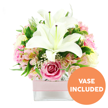 A delicately balanced pink and white floral arrangement rests against a plain white background, its soft colours and precise structure suggesting the handiwork of a skilled local florist in Mill Hill. Dominating the centre are striking white Oriental lilies, their petals broad and gently recurved, forming a sculptural star shape that draws the eye in to the dark reddish-brown stamens standing proudly in each bloom. Around these lilies, numerous soft pink roses are nestled, some still in tight buds with smooth, spiralling centres, others fully open with many layers of petals that create a lush, cushion-like texture. Threaded through the roses are stems of alstroemeria in gentle shades of blush and ivory, their slim petals enhanced by light yellow tones and fine speckles, adding delicate visual interest. Wispy sprays of white baby's breath are scattered throughout, introducing a sense of lightness and space, as though a soft breeze from Mill Hill Park were passing gently through the bouquet. Rich green foliage surrounds the flowers and peeks between them: shiny, broad leaves form a solid base, while lighter, feathery greens add movement and a fresh, garden-picked character. The entire composition is arranged in a modern, square clear glass vase, with the neatly trimmed stems visible through the water, emphasising the freshness of the design. A pale pink satin ribbon is wrapped horizontally around the middle of the vase, providing a subtle decorative touch without distracting from the blossoms. In the lower right corner of the image, a bright orange circular label with bold white capital text announces "VASE INCLUDED," making it easy to see that the container comes with the flowers. This bouquet feels serene yet celebratory, ideal for a bedside table in a Mill Hill home or as a charming gift delivered to mark birthdays, thank-yous, or new beginnings.