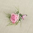 A delicate pink rose boutonniere is carefully laid on a softly textured, light beige linen surface, the fabric's natural weave forming a calm, neutral backdrop like a tablecloth in a Mill Hill townhouse near Mill Hill Broadway. Viewed from a gently elevated angle, the single rose is the clear focal point: its velvety petals are fully opened yet still neat at the centre, moving from a warm blush pink in the heart to a slightly deeper rose-magenta at the softly ruffled edges. Around the base of this romantic bloom, airy sprigs of white baby's breath cluster like tiny clouds, each miniature flower adding a light, lacy texture that softens the design. Slender strands of fresh, grass-like greenery arch gracefully from behind the rose, their pale green tones and fine lines giving the piece an effortless, natural movement, reminiscent of the greenery in local Mill Hill parks after spring rain. A broader, darker green leaf is folded and wrapped neatly around the base of the stems, giving the boutonniere structure and a polished, tailored look that would sit perfectly on a suit lapel for a wedding, prom, or black-tie dinner on The Ridgeway. The stems are bound in a narrow pink satin ribbon, tied into a small, precise bow that echoes the rose's colour and adds a gentle sheen where the light falls across it. Soft, even lighting casts subtle shadows that model the petals and foliage, enhancing the three-dimensional shape without overwhelming the scene. The overall impression is of quiet romance and expert craftsmanship, a small yet meaningful floral detail ready to be pinned to the jacket of a groom, groomsman, or special guest for a day of celebration in Mill Hill.
