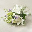 Set on a soft, off-white linen background that echoes the gentle charm of Mill Hill homes, this horizontal hand-tied bouquet radiates fresh sophistication. The centrepiece features grand white Oriental lilies-some fully open, revealing glistening petals and vibrant yellow anthers, while others gently unfurl, caught in a moment of quiet transformation. Delicate lisianthus, their petals crisp white and tipped with violet, bring a touch of playfulness and depth, their buds peeking through in anticipation of new beginnings. Threading upwards, slender purple veronica blooms add vertical texture, much like the native flowers found near the paths of Mill Hill Park. Wisps of baby's breath float within the arrangement, lending a soft, cloud-like finish that feels as light and celebratory as a new promise. Framing the bouquet, rich green leaves and airy asparagus fern unravel in layers, mimicking the lush, rolling green spaces that define Mill Hill. The stems are neatly bound with a white ribbon, ready for a ceremony or heartfelt delivery to a loved one. The interplay of crisp whites and deep purples feels modern yet timeless, making this bouquet an inspired choice for weddings, special occasions, or bringing a breath of North London's natural beauty into any setting.