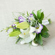 This arrangement reads as a study in soft contrasts and careful craftsmanship, arranged on a neutral, off-white linen surface that nods to Mill Hill's simple, classic interiors. A cluster of creamy white Oriental lilies anchors the bouquet: several blooms open to reveal arching, satiny petals marked with subtle dark speckles and prominent orange-yellow stamens dusted with pollen, while other buds remain tightly furled and vividly green. Interwoven are white-and-purple lisianthus, their ruffled faces graduating from a pale white centre to a lively lilac and deeper violet at the edges, petals that seem hand-painted and add a romantic, slightly whimsical flourish. Wisps of Baby's Breath float through the grouping, lending an airy, cloud-like lightness that balances the more statuesque lilies. The greenery is abundant and textural - glossy broad leaves like Salal or Ruscus, delicate feathery fronds and narrow grassy spikes give depth and a spectrum of greens that range from deep emerald to a soft, muted sage. The bouquet is finished with a neat white satin wrap, secured with small pearl-like accents that catch the light without ostentation. As a Mill Hill florist familiar with local weddings near The Broadway and small ceremonies at Mill Hill Old Church, I see this as an ideal bridal posy: fresh, elegant and quietly fragrant, with a faint spicy lily perfume and the sweet whisper of eustoma. Flowers Mill Hill prepares similar hand-tied bouquets for deliveries across Mill Hill and nearby Finchley, always aiming for tactile beauty and lasting freshness.