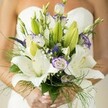 In the heart of Mill Hill, a bridal bouquet rests softly in the hands of an expectant bride-a composition that channels airy elegance and exuberant hope. Generous Oriental lilies glow with creamy white petals, subtly sprinkled and haloed by soft yellow stamens, filling the air with the sweet scent that often lingers amid Mill Hill's garden squares on spring mornings. Between the lilies, lisianthus blossoms open with fluttering, ruffled petals, each edged in a gentle sweep of lavender-as delicate as the afternoon light filtering through local chapel windows. Height and vibrance come from slender, deep purple Veronica, while baby's breath, airy and pure, weaves through the bouquet, imparting a dreamlike quality. The lush foliage-broad, dark-green lily leaves and airy fronds of asparagus fern-spills gracefully, echoing the rolling greenery of Arrandene Open Space nearby. The bride's palms are tenderly wrapped around the stems, her fingertips adorned with soft blush nail polish, the bouquet set against the gentle curves of her strapless wedding dress providing a pale, cloudlike background. This artfully gathered bouquet brims with artistry, anticipation, and a sense of new beginnings-an emblem of celebration handcrafted in Mill Hill, ready to grace weddings across NW7 with beauty and joy.