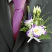A close-up wedding portrait focuses on the upper torso of a man dressed in elegant formalwear, standing as if ready to greet guests at a Mill Hill ceremony. His dark charcoal grey suit jacket is sharply tailored, the lapel smooth and slightly matte, framing a crisp, light grey checkered shirt beneath. At the collar sits a lustrous magenta silk tie, the knot perfectly formed, catching the light with a gentle sheen that adds vibrancy against the more muted fabric tones. Pinned neatly to the left lapel is a meticulously crafted boutonniere, clearly the work of an experienced local florist. At its heart are white lisianthus blooms edged in rich violet purple, their ruffled petals curving delicately around bright yellow centers. Around them cluster slender white buds, still closed and full of promise, interwoven with a slim, upright spike of deep indigo veronica that introduces height and a touch of wild-garden character. Wisps of feathery greenery and glossier emerald leaves cradle the flowers, some strands of fine decorative grass looping in soft arcs for a modern, fluid touch. Tiny white filler flowers, reminiscent of baby's breath, add a cloud-like softness and texture. A small white satin ribbon is tied into a precise bow at the base, securing the stems and lending a classic, bridal finish. The overall feel is quietly joyful and refined, like something you might see at a church wedding off Mill Hill Broadway or a celebration at a local country club, with the colours echoing romance and thoughtful attention to detail as the groom prepares for a day of lasting memories.