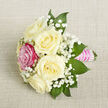 A delicately composed petite bouquet lies on a diagonally placed panel of light beige, finely textured fabric, reminiscent of woven linen or soft burlap, its neutral tone allowing every floral detail to stand out. At the centre of the design, five ivory to creamy white roses unfold in layered spirals, each petal smooth and velvety, giving an impression of calm elegance and quiet luxury. Just off to the front-left, one vivid fuchsia-pink rose takes pride of place, its richer colour drawing the eye immediately and offering a lively contrast to the serene whites. Between the roses, airy clusters of white baby's breath weave gently, their tiny rounded blooms forming a mist-like veil around the more substantial flowers. Surrounding the cluster, deep green, pointed leaves-likely ruscus or salal-provide a fresh, structured frame, their glossy surfaces echoing the well-tended greenery along the streets and gardens of Mill Hill. The stems are bound tightly together and wrapped in a bright pink satin ribbon that forms a neat handle, lying smoothly against the fabric backdrop. Over this satin base, several precise rows of small, iridescent white pearls are fixed in parallel bands, adding a refined, jewellery-like detail that catches the soft, even lighting. The composition feels intimate yet special, well suited for a corsage, boutonniere-style accent, or a small bridal posy carried at a Mill Hill ceremony, perhaps after photos on The Ridgeway. Crafted with care like those prepared daily at Flowers Mill Hill, the bouquet radiates romance and thoughtful celebration, perfect for weddings, proms, or meaningful anniversaries.