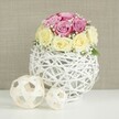 A serene floral display unfolds on a crisp white surface, evoking an intimate wedding table setting in Mill Hill, perhaps overlooking the green spaces near Mill Hill Park. At the heart of the scene is a rounded bouquet of roses, around a dozen blooms arranged in a tight, balanced dome. The colour palette is soft and romantic: half the roses are a gentle lavender-pink or fuchsia tone, while the others show creamy, buttery yellow petals that transition into pale ivory at their tips. Each bloom reveals delicate layers-some petals forming neat, central whorls, others flaring outward into fuller, more open shapes. Laced among them are airy tufts of white baby's breath, their tiny blossom clusters creating a mist-like texture that fills the spaces between roses and adds a light, bridal touch. The flowers nestle into a distinctive spherical vase crafted from slender, white-painted wicker or twig-like strands, woven into an open lattice. This intricate globe feels both rustic and contemporary, allowing glimpses of the stems and inner structure and casting gentle shadows on the white tabletop below. To the left, two pale rattan balls sit casually yet deliberately on the same surface, one slightly larger than the other, echoing the woven pattern of the vase and bringing harmony to the arrangement. Behind, a neutral beige or light taupe wall with a soft, fabric-like texture sets a calm, unobtrusive backdrop. The lighting is soft and even, with no harsh contrasts, enhancing the gentle colours and curves of the petals. The overall mood is one of quiet, refined celebration, ideal for illustrating wedding table arrangements, anniversary dinners, or sophisticated home decor curated for Mill Hill residents.