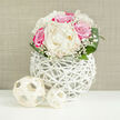 This image captures a refined floral display that could easily be imagined in a Mill Hill townhouse, arranged as a focal point for a wedding breakfast or anniversary dinner. The bouquet itself is compact and domed, shaped like a classic bridal bouquet resting gracefully in its stand. Dominating the centre are large creamy white peonies, their petals soft, plentiful, and gently undulating, each bloom opening into a full, almost pillow-like form. Encircling them are clusters of vibrant fuchsia-pink roses, the colour rich and saturated, with each petal curling inward to form a tight, elegant spiral. The contrast between the creamy whites and vivid pinks adds a romantic drama without feeling overpowering. Fine sprigs of white baby's breath thread their way through the arrangement, casting a lace-like network of tiny flowers that lighten the overall look and introduce a sense of air and space around the heavier blooms. At the base, smooth, deep green leaves arch outward, their surfaces subtly glossy, giving a polished finish and grounding the design. The flowers are held in a unique white vase constructed from interwoven, twig-like strands, forming a spherical, basket-style lattice. This open structure allows light to pass through, revealing glimpses of stems and shadow and creating delicate patterns. On the bright white tabletop in front of the vase rest two small decorative spheres, also woven and pale, their sizes slightly different but their textures echoing the vase. The background is a soft beige, possibly fabric or textured wallpaper, evenly lit so there are no harsh shadows. The complete scene evokes understated elegance and warmth, well suited to wedding flowers delivered around Mill Hill Broadway, reception décor in local venues, or stylish home arrangements that bring a touch of romance to everyday life.