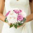 This bouquet exudes a captivating tranquility, the kind of calm beauty you might feel walking along the quiet residential streets of Mill Hill just before a wedding ceremony. The arrangement is neatly gathered, forming a rounded, hand-tied design that looks both polished and personal. At its heart are grand, creamy peonies, their petals unfurling like a soft whisper. Each bloom is densely layered yet airy, displaying a velvety texture that almost seems to invite your fingertips. Interwoven among the peonies are roses in dusty pink and soft violet hues, their tones subtle yet warm, creating gentle ripples of colour throughout the bouquet. These roses appear freshly gathered, their spiralling petals hinting at the promise of fuller opening as the day unfolds. Wisps of gypsophila peek delicately between the larger flowers, like tiny white stars dotting a pastel sky, adding a classic bridal charm that feels timeless in a North London setting. Shiny green leaves encircle the base, forming a natural cocoon around the blooms, enhancing their colours and adding depth. The lighting is soft, as though the bouquet is resting near a window in a Mill Hill home, the daylight catching the curves of each petal. You can almost sense a gentle fragrance blending notes of peony sweetness and rose warmth. The overall impression is intimate and refined, a bouquet that looks as though it has been thoughtfully created by Flowers Mill Hill for an elegant wedding where every detail is meant to echo love, harmony, and grace, perfectly suited to a ceremony at a local church or a reception on a leafy estate.