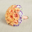 Captured from above on a neutral, textured fabric backdrop, this rounded bouquet presents a harmonious study in peach and violet nuances that speaks of understated elegance. The composition centres on abundant peach roses-each petal layered like a soft, unfolding promise-forming a compact, sumptuous dome that feels tactile and richly fragrant. Interlaced among the roses, white lisianthus with their violet-tinged edges introduce a cool, unexpected note, their ruffled profiles creating pockets of shadow and depth that make the warmer roses read even more luminous. Sprays of delicate white baby's breath weave between the larger blooms, providing a featherlight contrast and an impression of breath and movement. The bouquet's handle, shown extending toward the lower right, is carefully bound in a satin ribbon of coral-peach which gathers the stems into a reassuring, smooth embrace; a string of cream pearls spirals down the ribbon, adding an old-world gleam and tactile detail that catches the eye. The neutral linen surface and soft side light flatten into a calm palette, letting petals' textures and subtle colour gradients become the focal point-ideal for a refinement-seeking couple planning a Mill Hill Village celebration or a nearby church ceremony. Scent, too, seems present: the warm, honeyed rose aroma tempered by the fresh, green lift of lisianthus and the whisper of gypsophila. This bouquet offers both classic romance and contemporary flair, a quiet yet confident choice for anniversaries, intimate weddings, or keepsake photographs taken near Mill Hill Park.