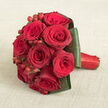 A lush cluster of deep red roses forms the heart of this bouquet, resting elegantly on a pale beige, textured fabric that recalls natural linen laid across a dining table. The arrangement is tightly composed into a rounded, almost domed shape, the roses pressed closely together so their velvety petals overlap in a seamless sea of crimson and dark burgundy. Each flower shows distinct character: some are wide open, revealing intricate, spiralling centres, while others are caught just at the moment of opening, their outer petals slightly curled and full of promise. Tucked between the rose heads are neat sprigs of red hypericum berries, their small, glossy orbs catching the light and adding a rhythmic pattern of shine and texture. Encircling the blooms, broad dark green leaves are folded around the base in a smooth collar, lending structure and a refined, modern edge that sets off the intensity of the red. Their slightly reflective surface contrasts gently with the matte background and the plush petals above. The bouquet's handle, visible to one side, is wrapped in vibrant red satin ribbon, wound so neatly that no stems are visible, creating a polished finish that would feel firm yet smooth in the hand. The viewpoint is slightly elevated, as though you are standing beside a table in a Mill Hill home, considering this bouquet for a special occasion. The image feels ideal for a romantic wedding at a local venue or a surprise anniversary gift delivered around Mill Hill East and Hendon, where the timeless symbolism of red roses still speaks louder than any written message.