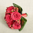 Viewed from above, this intimate hand-tied bouquet lies on a pale beige fabric background, its gentle weave and neutral tone allowing the flowers to command attention much like a carefully arranged centrepiece on a Mill Hill dining table. At the centre, five or six deep red roses cluster closely together; each bloom is in full flower, with satin-smooth petals spiralling toward a deep, romantic core that feels both lush and deliberate. Weaving through the roses are small sprays of red hypericum berries, their round, polished forms and slightly brighter hue adding a lively, textural counterpoint, as if scattered embers of light were dancing among the petals. Surrounding the blooms, large tropical leaves in rich, dark green have been shaped and curled to create a protective frame, their smooth surfaces and strong lines lending a modern, almost architectural character that gives the bouquet a contemporary flair well suited to Mill Hill's blend of leafy calm and urban style. At the base, a red satin ribbon wraps the stems in a compact band, adding both comfort for the hand and a coordinated finishing touch that echoes the roses' depth of colour. Soft, diffused lighting washes over the bouquet, highlighting the interplay of matte petals, glossy berries, and reflective leaves without any harsh glare, as though it were photographed near one of Mill Hill's quiet residential windows on a cloudy afternoon. The arrangement radiates feelings of affection, celebration, and poised romance, ideal for weddings, milestone anniversaries, or a meaningful delivery to a home near Mill Hill Broadway.