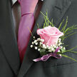 Pinned neatly to a dark, charcoal-grey lapel, this pink rose boutonniere offers a moment of softness and romance against the formal backdrop of a suit, as if ready to be worn at a wedding reception overlooking the green spaces of Mill Hill. The rose at the centre is a clear, gentle pink, its petals unfolding in even, velvety layers that create a rounded, perfectly formed bloom. Each petal catches the light along its edge, giving the flower a subtle glow, like the warm sky at sunset over Mill Hill Park. Surrounding the rose are slender fronds of feathery greenery, reaching upward in fine lines that add a sense of movement and structure. Tucked between these green accents, clusters of tiny white gypsophila flowers create a cloud-like border, their miniature blossoms bringing an air of innocence and celebration. The interplay between the delicate petals, the crisp green needles, and the matte texture of the suit fabric produces a balanced, sophisticated composition. At the base of the arrangement, a satin lilac ribbon is tightly wrapped and tied, its smooth surface reflecting just enough light to add a polished, ceremonial note. The ribbon's colour gently bridges the rose's blush tones and the snowy gypsophila, tying the palette together the way coordinated wedding flowers might be styled throughout a Mill Hill venue. One can almost sense the subtle fragrance of fresh rose mingling with clean, green notes from the foliage, creating an invisible aura around the wearer. This boutonniere feels like a handcrafted expression of joy and respect, a small but significant floral emblem for a groom, best man, or beloved guest in the heart of Mill Hill's community celebrations.