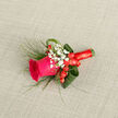 Resting diagonally across a pale, linen-like fabric, this boutonniere reads as a study in texture and colour harmony, the composition whispering of careful floral craft in Mill Hill. The central element is a single, lively magenta-pink rose whose petals curl inward at the centre and open outward in a graceful spiral, suggesting a bloom at the cusp of full expression. Around it, tiny clusters of white gypsophila float like miniature clouds, imparting an airy softness that offsets the rose's velvety intensity. Here and there, bright red hypericum berries introduce punctuated gleams of crimson, small globes that add festive cheer and a tactile contrast to the soft florals. The greenery is deliberate and varied: slender, wispy grass-like fronds provide movement, while broader, glossy leaves-carefully folded and tucked-lend sculptural structure and depth. The bouquet's base is wrapped in a bright red satin ribbon, wound with precision to secure the stems and to contribute a final flash of colour that ties the piece together. Envisioned for a prom night, a wedding procession along Mill Hill's tree-lined streets, or an anniversary dinner near Mill Hill Village, this boutonniere suggests a gentle fragrance of rose over fresh foliage and leaves a quiet, elegant impression - an intimate accessory designed to be both seen up close and remembered.