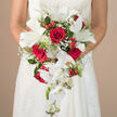 In this image, the camera focuses closely on a dramatic cascading wedding bouquet held at waist level by a bride in Mill Hill, her white gown detailed with fine lace or embroidery that adds a subtle texture around her arms and bodice. The background has been softened into a neutral, creamy blur, similar to the gentle tones you might see in the historic buildings and leafy streets near Mill Hill Broadway, allowing the bouquet to become the vivid focal point. Deep crimson roses form the heart of the design, their petals tightly layered yet fully open, creating velvety spirals of colour that feel rich and romantic. Between these roses, elegant white lilies stretch out wide petals, revealing pale green centres and visible stamens, adding structure and a fresh, almost luminous quality. The bouquet takes on its signature teardrop appearance through trailing white Phalaenopsis orchids that cascade downward from the main cluster. Each orchid stem shows open blooms and unopened buds arranged in sequence, their waxy petals catching light and giving a graceful, flowing line to the composition. Throughout the bouquet, tiny white flowers of baby's breath create a mist-like softness, weaving in and out of the larger blooms and softening the contrast between red and white. Glossy red hypericum berries add small, cheerful bursts of colour and texture, peeking out between leaves and petals. The greenery-ranging from slender, fern-like fronds to more substantial, polished leaves-frames and supports the flowers, enhancing both depth and movement. Crafted with the care of an experienced local florist, perhaps from Flowers Mill Hill, this bouquet feels suited to an elegant North London wedding, carried from a quiet residential street to a nearby church or country hotel, radiating love, freshness, and quiet sophistication.