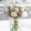 A tender sympathy bouquet, composed with quiet care, is the central focus of this image, evoking the understated elegance often found in Mill Hill's homes and chapels. The arrangement features a gathering of creamy white roses, each in full bloom, their petals gently unfurling in exquisitely layered spirals that create soft, rounded shapes. The roses' colour leans toward warm white, with subtle ivory undertones, lending them a comforting depth rather than stark brightness. Between and around these central blooms, clusters of tiny lavender and pale pink filler flowers form a gentle haze, their small, papery florets catching the light in a way that gives the bouquet an airy, almost ethereal quality. Together, the larger roses and the delicate filler blossoms strike a balance between strength and fragility, much like the emotions carried in a condolence message. All the stems are gathered into a tall, cylindrical clear glass vase filled with fresh water, allowing the eye to follow the straight green stems as they descend and lightly crisscross below the waterline. The vase rests on a smooth, white surface that quietly reflects the arrangement, while the background dissolves into a soft blur of pale tones, hinting at a serene interior setting somewhere near Mill Hill Broadway or along the quiet residential roads close to Mill Hill Park. The lighting is gentle, without harsh shadows, creating a tranquil mood. The image as a whole conveys a sense of stillness and compassion-a bouquet thoughtfully suited to sympathy deliveries in Mill Hill, meant to sit quietly beside a framed photograph or a flickering candle, offering a silent expression of support and remembrance.