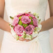 This bridal bouquet rests gracefully against a flowing wedding dress, its colours and textures coming together like a calm breath before the ceremony in Mill Hill. At its heart, a cluster of six full roses in a muted lilac-dusty pink hue steals the eye, each bloom richly layered with velvety petals that curl gently outward in perfect symmetry. Dotted amongst them, lively lime-green button chrysanthemums peep through, their rounded, finely textured heads bringing a spark of freshness and an easy sense of modern style. The entire arrangement is framed by a delicate border of creamy white flowers and slender buds, forming a soft, lace-like collar that mirrors the kind of filigree detail you might spot on bridal gowns in local Mill Hill boutiques. The bouquet is tightly hand-tied, the stems aligned and wrapped in a smooth ribbon so that only a hint of green peeks out at the base. In the soft natural light you might imagine filtering through a window overlooking Mill Hill Park, the colours appear gentle yet defined, with shadows resting lightly between petals. The scent is subtle but present: rose sweetness laced with cool greenery, quietly uplifting and soothing to nervous hands. There is a lovely balance between structure and spontaneity, as if the flowers were chosen carefully from a trusted florist, yet gathered with an easy, garden-like touch. This bouquet feels designed to move with the bride-from first photos near Mill Hill Broadway station to evening celebrations-remaining elegant, fresh, and utterly in tune with the emotion of the day.