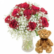 A serene, high-contrast photograph presents a classic romantic gift set likely to be found in a Mill Hill flower shop: an elegant red rose arrangement paired with a cuddly teddy bear, both highlighted against a crisp white background. On the left, a clear glass vase rises from the white surface, holding about a dozen deep crimson roses. The blooms are lush and full, with velvety petals swirling gracefully around their centres, some just beginning to open while others are expanded into generous rosettes. Threads of delicate white baby's breath weave between the roses, their tiny blossoms forming a soft, cloud-like surround that lightens the intensity of the red, much like pockets of sunlight filtering through trees in Mill Hill Park. Dark green foliage and stems provide grounding contrast, and through the transparent vase you can see the straight stems submerged in crystal-clear water, suggesting a freshly crafted bouquet prepared for local delivery. To the right, a medium-sized teddy bear sits upright, turned slightly toward the flowers as though keeping them company. Its fur is a gentle light brown plush, with a soft, slightly shaggy texture that seems made for hugging. The bear's details are simple yet expressive: rounded ears, shiny dark eyes that catch the light, a small embroidered nose, and a faint stitched smile that feels kind and welcoming. A dark brown ribbon is tied around its neck, its satin texture catching a subtle sheen. The minimal, white background places all focus on this pairing of rich roses and soft toy, evoking the feeling of sending a heartfelt message across Mill Hill-ideal for anniversaries, romantic birthdays, Valentine's Day surprises, or quiet gestures of gratitude and affection.