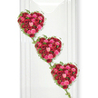 The photograph shows three sumptuous, heart-shaped arrangements of roses, designed as a cascading funeral tribute with a romantic yet dignified character, much like the quiet charm of Mill Hill's tree-lined avenues. The hearts are arranged vertically, gently offset in a diagonal line, so that they seem to flow down the image in a continuous expression of love and remembrance. Each heart is densely packed with roses in deep, velvety reds and vivid pinks, accented by occasional blush-toned petals that soften the palette. The roses are nestled tightly together, some in bud, others fully open, creating a richly textured surface where highlights and shadows play among the layered petals. Around the edge of each heart, fine ivy vines curl gracefully, their small, dark green leaves giving a clear outline to the shape before trailing down to join one heart to the next. This linking greenery adds movement and symbolism, evoking connection, continuity, and the bonds between family members, partners, or close friends. The arrangement is photographed against a bright white background that suggests a calm interior space, perhaps in a Mill Hill chapel or a dedicated room set aside for reflection before a service. The stark simplicity of the backdrop makes the colours of the roses appear more vivid and the greens more intense, while soft, diffused shadows give the hearts a sculptural presence. The overall effect is tender and heartfelt, an expertly crafted tribute that speaks of devotion, gratitude, and the wish to say goodbye with beauty and care, as might be created by Flowers Mill Hill for a local family farewell.
