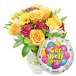 Against a crisp white backdrop, this 'Get Well Soon' gift ensemble feels like sunshine wrapped in glass. On the left a generous bouquet sits in a clear glass vase; stems and cool water are visible beneath a tight spray of glossy green foliage that cradles the blooms. The composition centres on soft, opening roses in warm orange and deep golden-yellow tones, their velvety petals unfurling in concentric spirals that suggest comfort and gentle optimism. Bright yellow gerbera daisies punctuate the arrangement with their broad, cheerful faces, while ruffled carnations in a rich burgundy or maroon add tactile depth and a comforting, plush contrast. Scattered between these are small, starry white alstroemeria (Peruvian lilies) offering delicate points of light and an airy balance to the denser blossoms. The whole bouquet gives off a faint, citrus-rose scent under soft daylight, a soothing fragrance suitable for a bedside or hospital room. To the lower right, partially overlapping the flowers, a round silver Mylar foil balloon reflects light with a silvery sheen; its centre shows a playful cluster of cartoon-style balloons in blue, purple, green, pink and yellow, some striped, some polka-dotted, and bold yellow-and-blue lettering with a discreet shadow reads Get Well Soon. Tiny starburst graphics halo the balloon's white border, and a small KALEIDOSCOPE logo sits at the very bottom edge. As a local Mill Hill florist might say, this pairing of bright blooms and a cheerful foil balloon sends an unmistakable message of warmth, encouragement and well-wishing, perfect for offering comfort to someone recovering at home or in local care near Mill Hill Broadway.