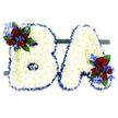 A tender, meticulously crafted funeral tribute shaped into the capital letters B and A, each letter built from hundreds of small, pristine white chrysanthemum florets that create a soft, pillowy surface. The chrysanthemums are layered so densely the letters read as cloud-white mosaics, their tiny petals catching a cool, diffused studio light that gives the whole piece a quiet, luminous calm. Each letter's silhouette is crisply defined by a ruffled royal-blue satin ribbon wrapped around the edges, the deep blue providing a solemn, elegant frame against the pure white face.
On the upper left curve of the B sits a compact spray of velvety deep-red roses, buds and half-open blooms mingling with spiky, electric-blue delphinium-like florets; similar accents appear at the lower right of the A, balancing the composition. Dark green leaves and paler trailing tendrils weave through the clusters, softening hard lines and adding a natural, living contrast to the formal letters. Both initials rest side-by-side on a slim, dark-green rectangular base that anchors the display, suggesting a freestanding memorial meant for placement at a service or outside a chapel. The spare white background keeps attention on texture and colour - the cool whites, royal blue trim, passionate reds and cobalt touches - a palette that reads as respectful, heartfelt, and quietly patriotic for some families.
As a local Mill Hill florist might explain to a grieving family, this arrangement speaks of clarity and remembrance: each bloom chosen for lasting shape and clean tone, suitable for viewing near Mill Hill Broadway or a funeral held close to Mill Hill Park, offering a composed, personal goodbye.