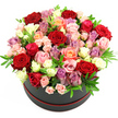The photograph presents a striking, dome-shaped bouquet of roses overflowing from a dark hatbox, styled in a way that suggests both luxury and modern romance. The hatbox itself is a deep charcoal or black tone with a soft, matte surface, ringed at its base by a slender, contrasting red band that adds a refined splash of colour. Above this simple cylinder, the flowers rise in a full, rounded mass, so densely packed that they form a continuous carpet of petals. At the core are large, velvety red roses with tightly layered centres, conveying passion and devotion, much like a romantic evening enjoyed in one of Mill Hill's cosy restaurants near the Broadway. Surrounding these are roses in gentle light pinks, some appearing as spray roses with multiple small blooms per stem, and others as standard blooms with more open faces. Peach and coral tones introduce a soft, warm glow, while creamy white flowers provide brightness and visual rest. Scattered among the pastels, delicate touches of lavender and light purple blooms add depth and a contemporary flair, reminiscent of the cool, misty mornings over Mill Hill's green spaces. Threads of lush green foliage, likely ruscus or eucalyptus, are tucked judiciously throughout, offering refreshing green accents that help define the shapes of the roses. The whole composition is lit evenly, with no harsh shadows, so every curve and ruffle of the petals is clearly visible against the plain white backdrop. The result is a bouquet that radiates affection, celebration, and thoughtful craftsmanship-an ideal floral gift for anniversaries, birthdays, Valentine's Day, or heartfelt thank-you moments delivered across Mill Hill's homes and businesses.