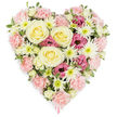 A beautifully detailed heart-shaped floral arrangement is pictured against a pure white background, glowing with soft tones that would feel perfectly at home in a quiet Mill Hill chapel or family living room during a time of mourning. The heart is tightly packed with blossoms so that they form a complete, unbroken surface of colour and texture. At its centre, several large roses in creamy ivory to pale yellow unfold in classic spirals, their velvety petals catching the light and symbolising warmth and devotion. Around these focal roses, an abundance of pastel pink carnations gathers, their densely ruffled petals giving the heart its plush, cushioned look and clearly tracing the gentle curves of the design. Threaded among the carnations are crisp white spider chrysanthemums, their long, fine petals radiating out from green centres like bursts of light, adding airy contrast and delicate structure. Deeper magenta-pink lisianthus blooms are tucked between the paler flowers, their softly fluted, bell-shaped petals bringing a richer hue and a sense of emotional depth. Tiny hints of fresh green foliage and buds peek through, suggesting natural movement and freshness, as though the flowers had been arranged shortly after arriving from a local Mill Hill flower market. At the very tip of the heart, a soft pink ribbon slips subtly into view, indicating this is a completed sympathy or funeral tribute, ready for delivery to a service in Mill Hill or nearby. The overall impression is of a lovingly crafted floral heart, offering comfort, remembrance, and a visible expression of love in moments when words alone are not enough.