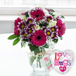 A warm, joyful arrangement is captured as if resting on a tidy table in a Mill Hill home, freshly delivered and still glistening with clarity from the vase water. The bouquet sits in a rounded clear glass vase, its sides revealing a fan of neatly cut green stems submerged in clean water. Above, the flowers form a generous dome. Large gerbera daisies in rich magenta and fuchsia tones bloom proudly at the front, their symmetrical petals encircling dark, velvety centres that add dramatic depth. Interspersed between them, smaller chrysanthemums offer layered texture and colour, their petals a gradient from deep purple at the base to crisp white at the tips, each bloom focused around a golden-yellow centre. Creamy white roses are tucked throughout the arrangement, some tightly furled, others slightly open, bringing a soft, romantic calm to the brighter hues. Threads of white baby's breath float between the larger flowers like a fine mist, adding delicacy and preventing the bouquet from feeling heavy. Surrounding foliage in fresh shades of green peeks from beneath and between the blossoms, shaping the bouquet into a full, rounded form reminiscent of the greenery seen along Mill Hill's quiet residential streets. Around the neck of the vase, rustic natural twine is tied in a relaxed bow, supporting a small white gift tag printed with a pink rose motif and gentle green script, suggesting a personal, thoughtful touch. To the lower right, partially overlapping the vase, a gleaming heart-shaped foil balloon with a silver edge adds a playful accent, its white centre emblazoned with the phrase Love n' Hugs 24-7 in a patchwork of pink, purple and blue letters. The softly blurred, bright background hints at natural daylight streaming through a Mill Hill window, creating an uplifting, affectionate mood perfect for marking special moments or simply brightening someone's day.