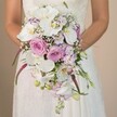 In this detailed wedding image, a Mill Hill bride is seen from her upper torso, hands resting gracefully around an extravagant cascading bouquet-its waterfall form a true testament to elegant local artistry. Her gown, a vision in soft ivory, features subtle lace that hints at traditional NW7 bridal style. At the bouquet's centre, snowy white Phalaenopsis orchids bloom with broad petals and sunbeam-yellow centres, their luxury harmonised by blush pink and lavender roses in all their fragrant fullness. Spheres of baby's breath add an airy, cloudlike touch reminiscent of garden mornings by Bittacy Hill, while smaller lavender blossoms and sprigs of stock or sweet pea provide depth and a play of soft purple hues. Darker, magenta amaranthus trails pour gracefully downward, their unusual texture creating movement and a slight sense of drama. Glossy green foliage and young buds peek amongst the flowers, giving the bouquet organic dimension and freshness. The soft, neutral background ensures the focus remains on the arrangement's pristine whites, romantic pinks, and serene purples-true to Mill Hill's refined wedding traditions. Lovingly crafted by Flowers Mill Hill's skilled florists, this bouquet is the perfect inspiration for couples seeking an elegant, modern touch for their local ceremony or reception.