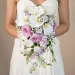Against a softly glowing taupe backdrop-a palette reminiscent of late afternoon sun in Mill Hill-a bride, pictured from mid-chest downward, presents a strikingly elegant cascading bouquet, its teardrop silhouette exuding sophistication and romance. The pristine white of the bride's strapless gown, adorned with delicate lacework, offers a subtle contrast to the lush, flowing floral display she holds. The bouquet's design centers on radiant phalaenopsis orchids whose broad ivory petals and yellow cores bring a serene opulence. Opulent pink garden roses burst alongside, each bloom adding a plush texture that suggests care and artistry honed in the heart of Mill Hill. Small lavender and mauve flowers-likely stock and lisianthus-intermingle, their hues and wispy shapes introducing depth, while flecks of white baby's breath thread through to soften the overall display. Sparse touches of green foliage punctuate the cascade without overwhelming it, maintaining a pastel-dominant palette. The bouquet's overall effect is one of classic, romantic grandeur-perfect for Mill Hill brides who seek both modern beauty and lasting tradition, brought to life by skilled local florists for weddings celebrated across NW7.