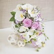 This image shows a sophisticated wedding-style table arrangement, crafted in a palette of white, soft pink, and gentle lavender that would feel perfectly at home in an elegant Mill Hill marquee or garden reception. The flowers are gathered into a distinctive white spherical vase whose open, woven design looks like a carefully sculpted rattan globe, its fine strands interlacing to create a sense of airy lightness. Resting on a spotless white surface, the vase is accompanied by two decorative cream-coloured woven balls, their lattice structure echoing the container and bringing the whole composition together. From this base, the bouquet rises in a lush, rounded form. Several large white Phalaenopsis orchids sweep across the top and sides, their broad, silky petals unfurling gracefully, each bloom centred by a hint of golden yellow that adds an understated touch of luxury. Nestled among the orchids are romantic roses in blush pink and muted lavender, some fully opened to reveal intricate spirals of petals, others just beginning to reveal their soft inner layers. Smaller, ruffled flowers in white and lilac with purple-edged petals are tucked between the larger blooms, adding depth and delicate colour variations. Fine sprays of white baby's breath weave through the arrangement, creating a misty, cloud-like veil that softens every angle. From the lower edge, slender stems of light purple blossoms spill gently downward, giving a sense of movement and hinting at a cascading effect. Fresh green foliage peeks out between the petals, framing the pastel tones and adding vibrancy. The softly textured beige background remains pleasantly out of focus, so the eye is drawn to the fresh, detailed flowers, an ideal visual for describing Mill Hill wedding tables, anniversary dinners, or refined celebration décor prepared by local florists.
