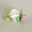 A delicate orchid boutonniere or corsage lies horizontally on a softly textured oat-coloured linen cloth, its pale beige background giving a calm, neutral stage that lets every floral detail stand out. At the heart of this handcrafted piece sits a single, flawless white Phalaenopsis orchid, its broad petals smooth and gently curved like folded silk. The centre of the orchid glows with soft yellow and the faintest blush of pink, adding warmth and depth to the otherwise pure white bloom. Around its base, tiny star-like blossoms of white baby's breath cluster together in airy clouds, creating a light, romantic halo that softens the lines of the design. Slender, fresh green leaves peek out from behind the flowers, adding a natural contrast and hinting at the living stem beneath. To one side, a short side-spray carries several unopened, pale lime-green orchid buds, their firm, closed forms suggesting new flowers ready to open later in the day. The stems are neatly gathered and wrapped with a slim, bright fuchsia satin ribbon, tied into a small, crisp bow whose sheen catches the gentle, even light. The vibrant pink ribbon offers a joyful pop of colour against the calm, neutral fabric and the white petals, giving the whole accessory a celebratory feel. The lighting is soft and diffused, like a quiet morning in Mill Hill before guests arrive for a wedding at a local church or a formal event in one of the nearby venues. The overall impression is of understated, timeless elegance: a carefully crafted boutonniere perfect for a groom, groomsman, or prom date, capturing both tenderness and tradition in a single, beautifully composed floral detail.