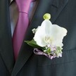 A close-up image shows a groom's charcoal suit in Mill Hill, the camera lingering on the lapel where a white Phalaenopsis orchid boutonniere rests in perfect balance with a rich plum tie. The suit jacket is expertly tailored, its deep, almost black fabric catching the light in soft, refined highlights along the clean edge of the left lapel. Against it, the smooth, lustrous tie in magenta-plum glows warmly, knotted neatly over a pale grey dress shirt with a discreet woven pattern that adds quiet texture rather than distraction. Pinned to the lapel, the orchid is the star: broad, velvety petals in pristine white open like a small sculpture, framing a vivid yellow and soft orange centre that draws the eye. Around the orchid, delicate clusters of baby's breath and tiny pale lilac blossoms gather like a floral halo, suggesting heather or similar woodland flowers you might find on an early walk through Mill Hill Park. Slender, fresh green leaves peek out, giving depth and a touch of natural wildness to the otherwise polished look. Just above the main bloom, a pale green orchid bud stands upright, hinting at the promise of new beginnings, much like a wedding day itself. At the base, a slim lavender satin ribbon is tied into an intricate bow, echoing the colour of the smaller flowers and the undertone of the tie, as if carefully coordinated by a local Mill Hill florist who understands both fashion and sentiment. The overall feeling is one of understated elegance and gentle romance, ideal for a groom or groomsman stepping into a ceremony in one of Mill Hill's churches or garden venues, ready for photographs that capture every small, meaningful detail.
