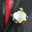 A close-up photograph shows an elegant wedding boutonniere pinned neatly to the lapel of a dark charcoal grey, almost black suit jacket, the fine wool fabric catching soft, natural light. The jacket's subtle woven texture frames the flowers, while to the left a vivid, solid red satin tie lies against a light grey and white small-checked dress shirt, adding a bold splash of colour that feels perfect for a Mill Hill celebration. At the heart of the boutonniere rests a single, large white Phalaenopsis orchid, its velvety petals gently arched and luminous, with a creamy yellow-and-white lip forming a sculptural centrepiece. Around the orchid, tiny white clouds of baby's breath cluster like soft mist, their miniature blooms delicate and airy, adding romance and lightness. Just above, two small, round lime-green chrysanthemum-style button blooms nestle in, their bright green hue and slightly tufted texture bringing a fresh, playful contrast to the serene orchid. A few rich green leaves cradle the composition, their smooth surfaces glinting slightly as they curve around the flowers, giving depth and a natural frame. The stems are tightly bound with a narrow deep red satin ribbon that subtly echoes the groom's tie, the sheen of the ribbon adding a refined, coordinated touch. The boutonniere is positioned just above the jacket's neatly stitched breast pocket, suggesting a groom ready to walk into a wedding at a Mill Hill church or a nearby venue on The Ridgeway, the flowers fresh and meticulously arranged, a quiet yet confident symbol of love and commitment captured in a single, thoughtfully designed detail.