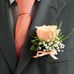 This close-up shows a peach-toned rose boutonnière arranged with intentional grace and pinned to the left lapel of a dark charcoal suit jacket, photographed in soft daylight that sculpts each petal and shadow. The rose is the quiet hero: petals gently unfurling in concentric layers, their surface velvety and slightly luminous. Nestled beside it, tiny white gypsophila provides an airy, cloud-like texture while fine, wispy green foliage offers delicate lines that lengthen the silhouette and catch the eye without overwhelming the bloom. A narrow satin ribbon in a pale peach-salmon hue is tied neatly at the base, its glossy fibres mirroring the sheen of the warm-toned tie worn over a light shirt patterned with a refined dark grid. The overall effect is one of handcrafted restraint - a boutonnière that balances softness and structure, ideal for a wedding or formal reception. As a Mill Hill florist familiar with local tastes, I picture this pinned during photos around Mill Hill Broadway or quietly fastened before a ceremony at a nearby chapel. The arrangement reads as familiar and modern at once, suitable for grooms who prefer subtle colour notes rather than loud florals. Flowers Mill Hill prepared pieces like this with attention to stem wiring, ribbon finishing, and fresh flower handling so they remain supple and fragrant through portraits and evening dancing, and we can advise on matching pocket squares or tie tones to complement the peach palette for deliveries across the Mill Hill area.